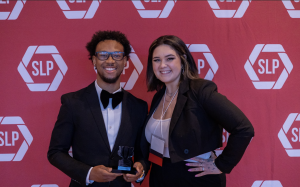 A man and a woman smiling in front of a backdrop holding an award.